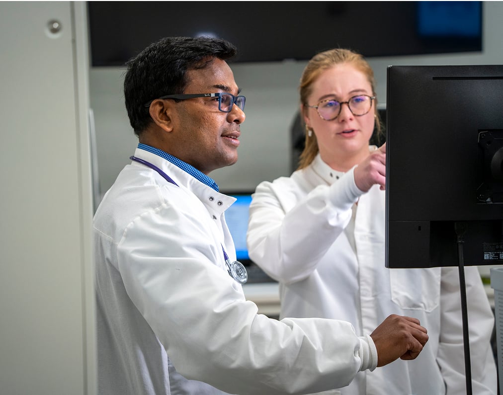 Two laboratory scientists in white lab coats collaborating while reviewing diagnostic results on a computer monitor.